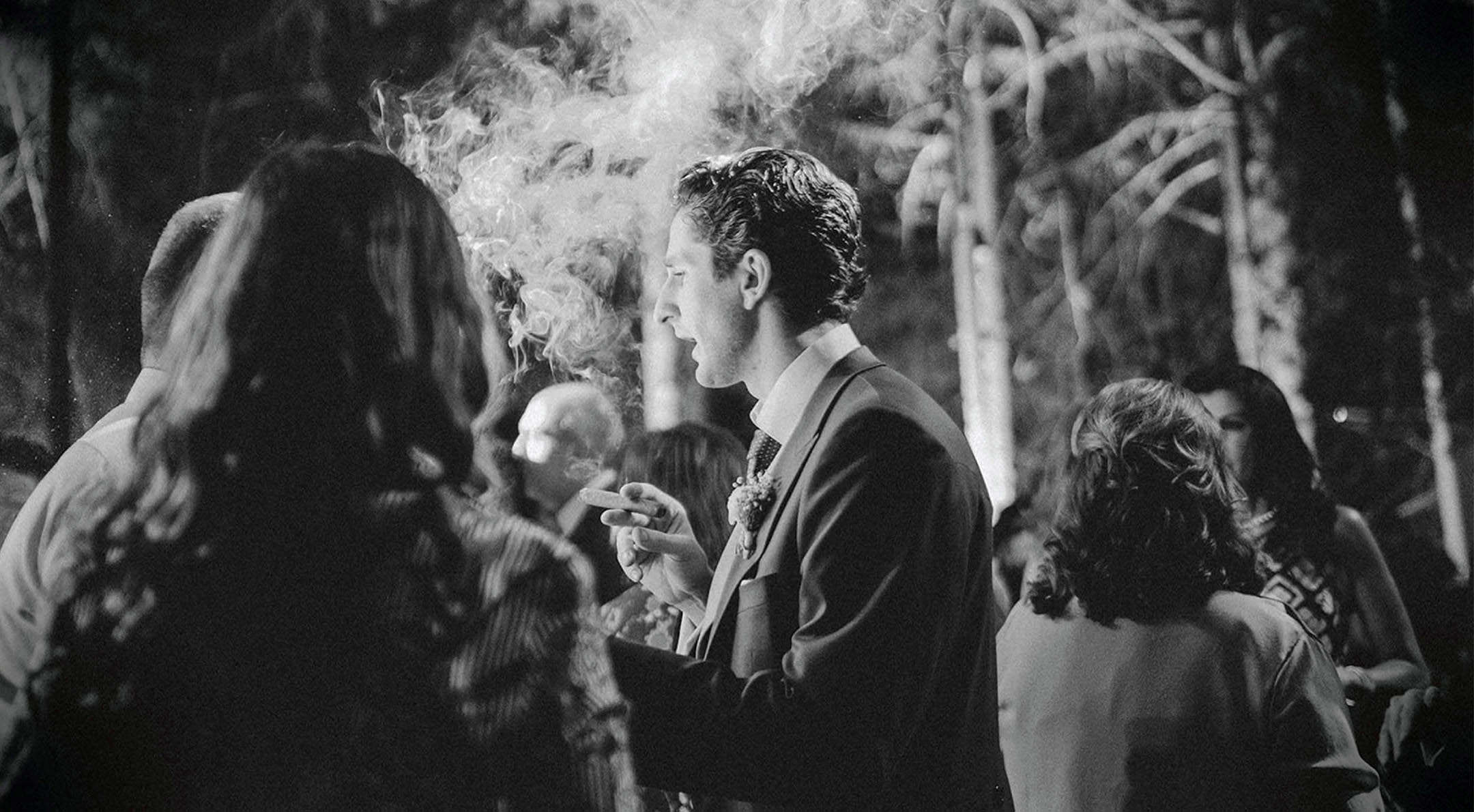 Breathless Weddings Groom exhaling smoke while talking during a nighttime wedding reception.