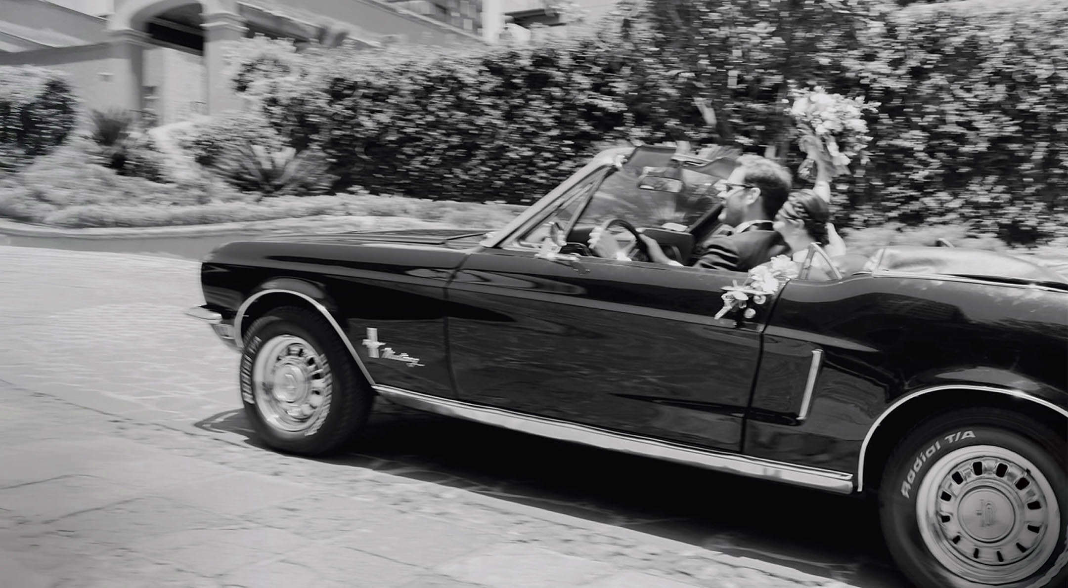 Breathless Weddings Bride and groom waving in a black vintage Mustang convertible